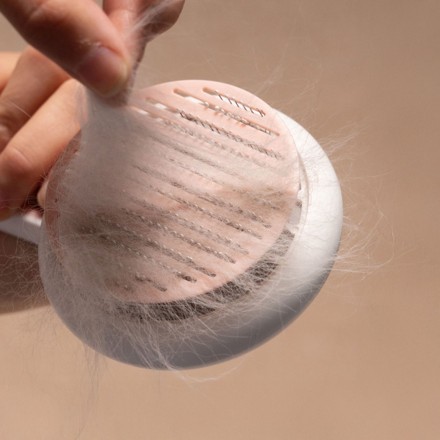 A hand peeling a thick layer of matted white fur off the surface of a self-cleaning pet grooming brush.